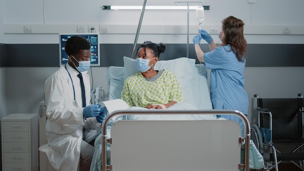 Kranker Patient, der medizinische Hilfe von einem Sanitäter mit Gesichtsmaske zum Schutz in der Krankenstation erhält. Arzt spricht mit Frau, während Assistentin während Pandemie mit Infusionsbeutel hilft.