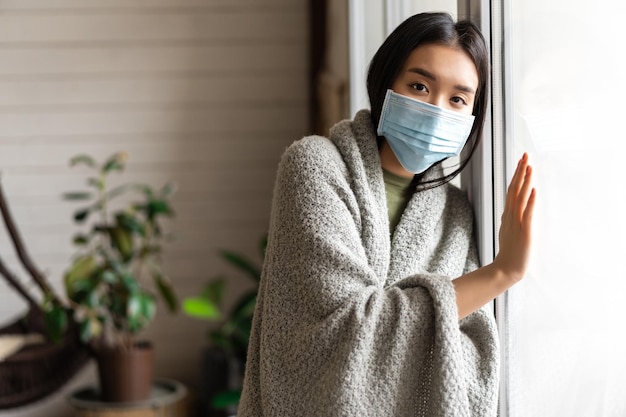 Kostenloses Foto krankes asiatisches mädchen in medizinischer gesichtsmaske, das am fenster steht und sich danach sehnt, nach draußen zu gehen, während es auf quar...