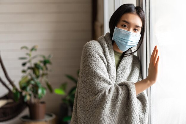 Krankes asiatisches Mädchen in medizinischer Gesichtsmaske, das am Fenster steht und sich danach sehnt, nach draußen zu gehen, während es auf Quar...