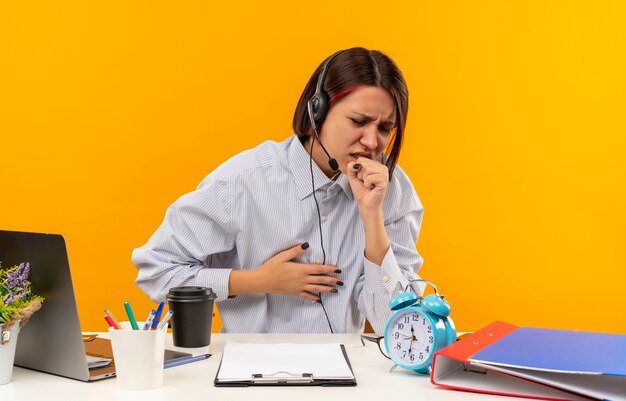 Krankes junges Callcenter-Mädchen, das ein Headset trägt, das am Schreibtisch sitzt, hustet und Hand auf Brust lokalisiert auf Orange setzt