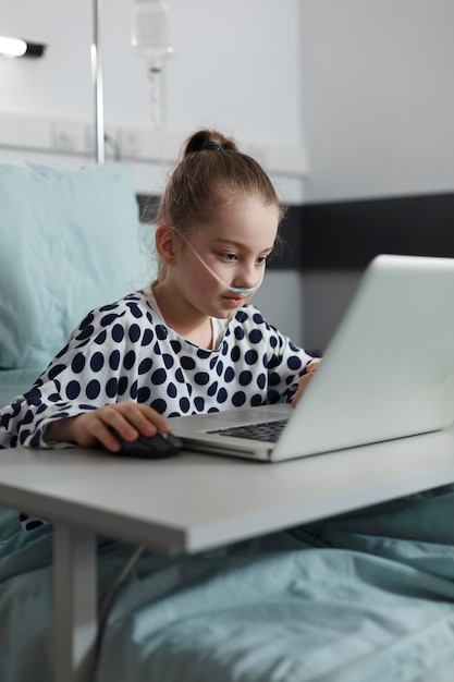 Krankes Kind, das sich auf dem Patientenbett ausruht, während es auf einem modernen Computer spielt. Krankes kleines Mädchen, das Spiele auf dem Laptop spielt, während es in der Kinderstation der Gesundheitsklinik stationär behandelt wird