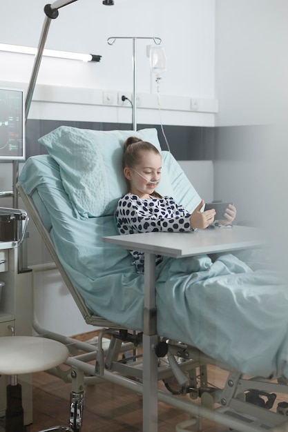 Krankes kleines Mädchen, das auf dem Smartphone spielt, während es sich auf dem Patientenbett im Behandlungsraum der Kinderklinik ausruht. Fröhlicher junger Patient, der Handy-Unterhaltung genießt, während er in der Klinik-Aufwachstation sitzt.