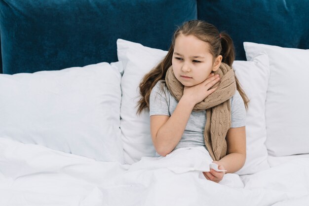 Krankes Mädchen, das auf Bett mit dem Schal um ihren Hals sitzt, der unter schmerzendem Hals leidet