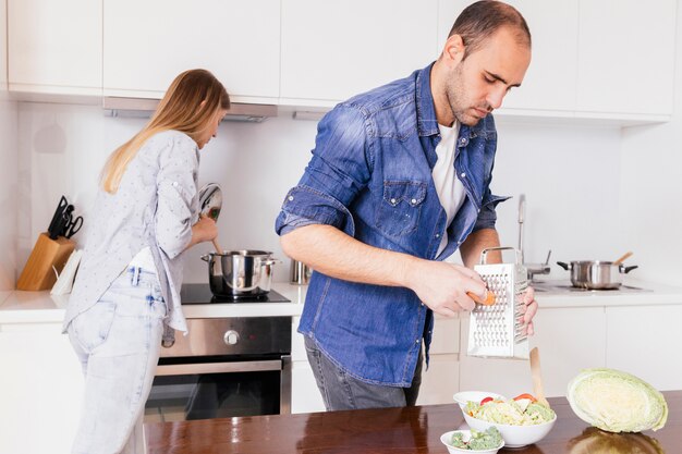 Kratzende Karotte des jungen Mannes mit seiner Frau, die Lebensmittel im Hintergrund zubereitet