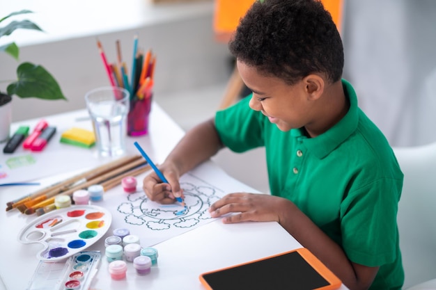 Kreative Aktivitäten. Profil eines dunkelhäutigen lächelnden Jungen in grüner T-Shirt-Zeichnung mit blauem Bleistift, der im hellen Raum am Tisch sitzt