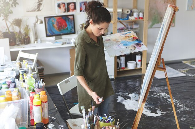 Kreative Frau, die Pinselstriche auf Staffelei macht, die in ihrer Werkstatt stehen, umgeben von verschiedenen bunten Ölen. Talentierter Maler, der Bild im Kunststudio mit Aquarellen und Pinsel zeichnet.