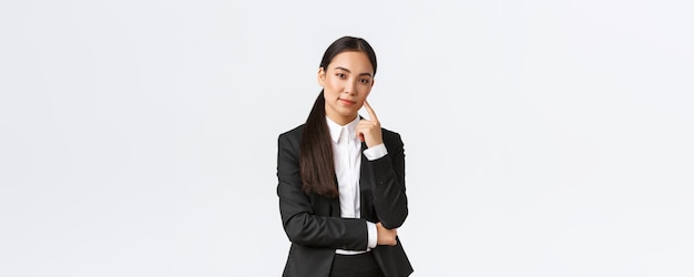 Kreative, intelligente asiatische Managerin im Anzug, die schlau lächelt und in die Kamera schaut, hat einen Plan, der denkt, dass sie eine ausgezeichnete Idee hat, die über weißem Hintergrund erfreut steht