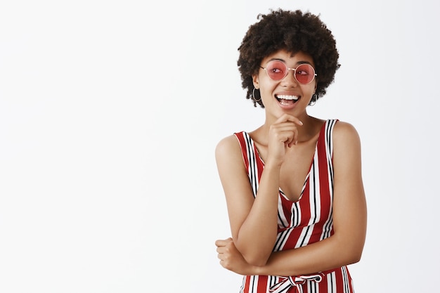 Kreative und stilvolle weibliche afroamerikanische Designerin in gestreiften trendigen Overalls und rosa Sonnenbrillen, die Hand am Kinn halten, lächelnd und mit Interesse nach links schauend