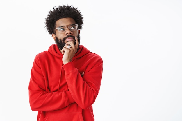 Kreativer afroamerikanischer bärtiger Typ mit Afro-Frisur in Brille und rotem Hoodie, der ein neues Lied kreiert, in nachdenklicher Pose steht und das Kinn berührt, das verträumt aussieht, auf die obere rechte Ecke konzentriert ist, denkend