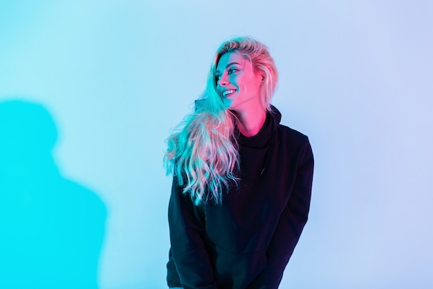 Kreatives glückliches porträt einer lächelnden schönen blonden frau in modischer kleidung mit schwarzem hoodie im studio auf mehrfarbigem rosa und blauem hintergrund