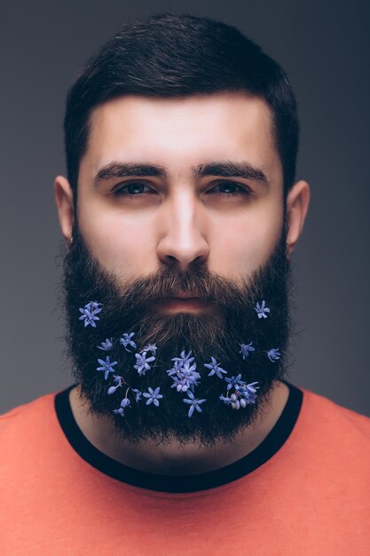 Kreatives Porträt des jungen schönen Mannes mit einem Bart, der mit Blumen verziert wird.