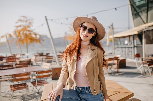 Kühlende junge Frau im lässigen Outfit, das durch Sonnenbrille mit Lächeln sitzt, das auf Tisch sitzt