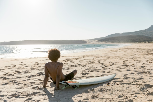 Kühler Mann, der auf Strand mit Surfbrett sitzt