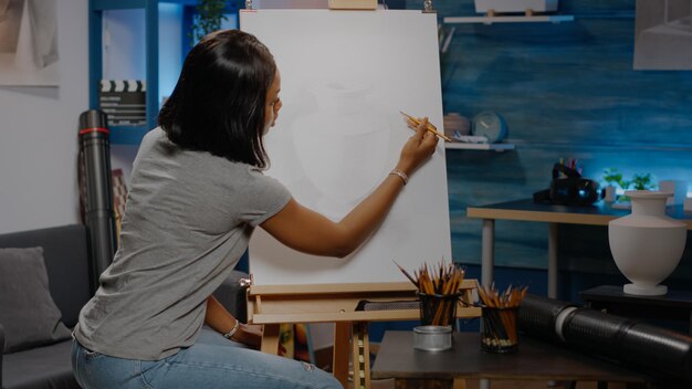 Künstler der afroamerikanischen Ethnizität zeichnet Vase auf weißer Leinwand mit Bleistift und Bastelwerkzeugen im Studio. Schwarze junge Frau, die authentisches Design für neue Meisterwerke und bildende Kunst kreiert