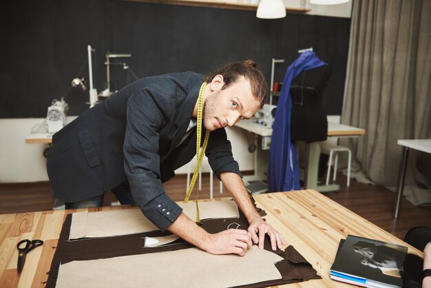 Künstler im Studio. Nahaufnahme des talentierten attraktiven jungen männlichen Modedesigners, der an der neuen Winterkollektion arbeitet, mit entspanntem Ausdruck in die Kamera schaut und Kleiderteile ausschneidet.