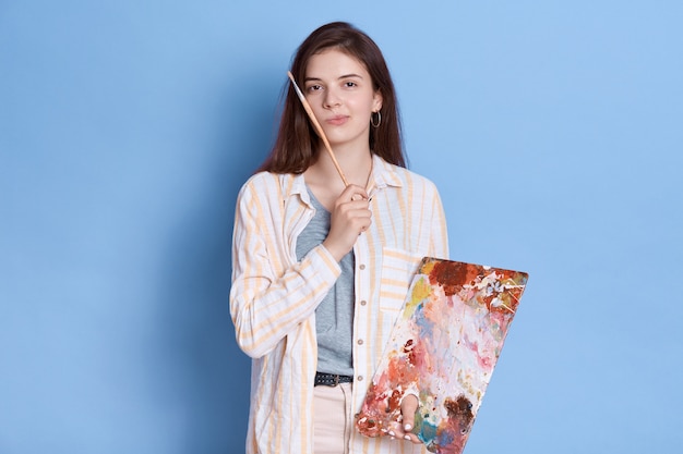Künstler malt Bild, posierend mit nachdenklichem Ausdruck, brünette Dame, die weißes Hemd mit Pinsel in den Händen trägt.