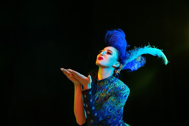 Küsse. Schöne junge Frau im Karneval, stilvolles Maskeradenkostüm mit Federn auf schwarzer Wand im Neonlicht. Copyspace für Anzeige. Feiertagsfeier, Tanz, Mode. Festliche Zeit, Party.