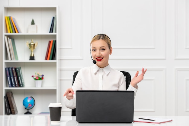 Kundendienst nettes blondes Mädchenbürohemd mit dem Kopfhörer und dem Computer, die auf papercup zeigen