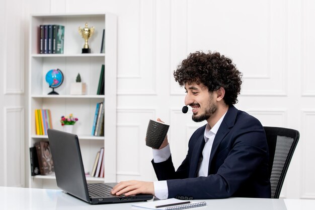 Kundenservice gutaussehender, lockiger Mann im Büroanzug mit Computer und Kopfhörer, der Kaffee trinkt