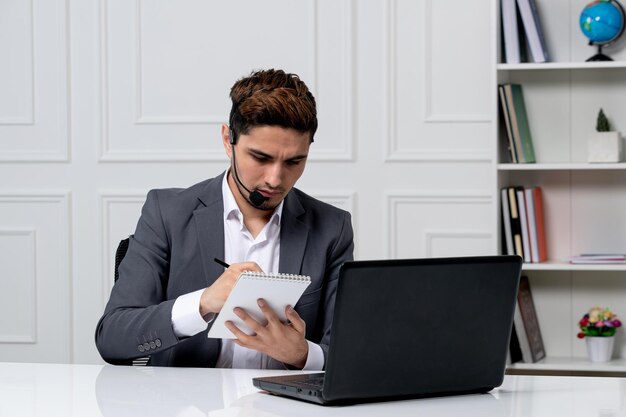 Kundenservice junger süßer Kerl im grauen Büroanzug mit Computer, der sich Notizen macht, fokussiert