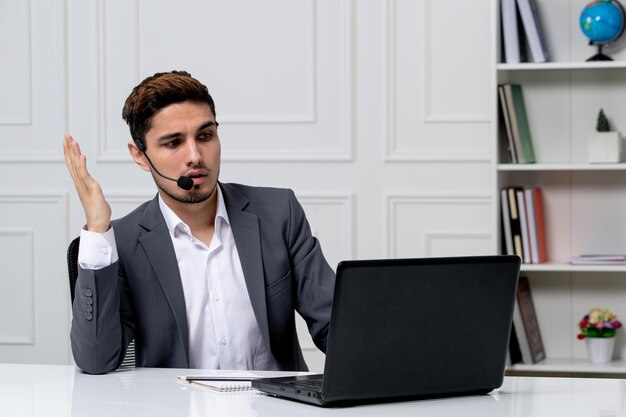 Kundenservice junger süßer Kerl im grauen Büroanzug mit Computer winkenden Händen während des Anrufs