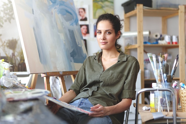 Kunst, Arbeit, Inspiration und Kreativität. Porträt der schönen talentierten jungen brünetten Künstlerin in Jeans und Hemd der khakifarbenen Farbe, die in ihrer Werkstatt vor Leinwand sitzt und am Malen arbeitet,