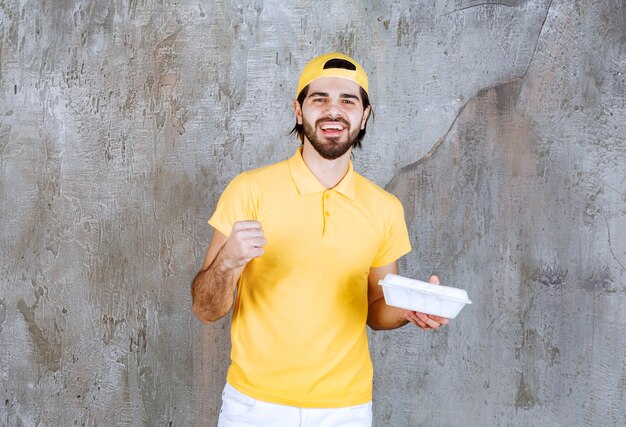 Kurier in gelber Uniform, der eine Plastikbox zum Mitnehmen liefert und ein positives Handzeichen zeigt