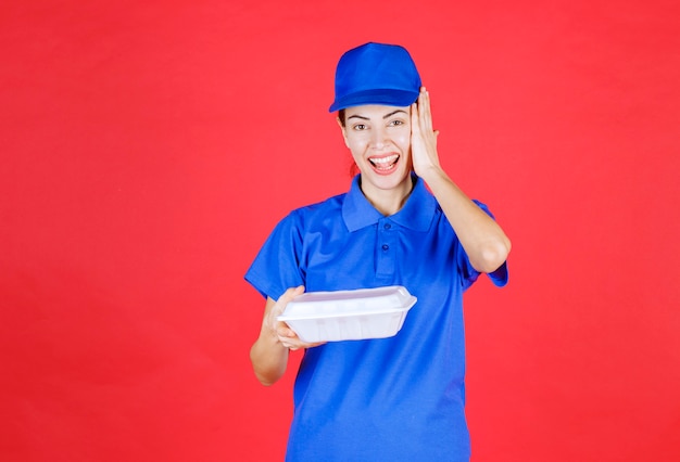 Kurierfrau in blauer uniform, die eine weiße box zum mitnehmen hält und sieht verwirrt und überrascht aus.