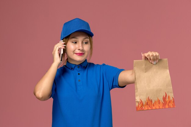 Kurierin in blauer Uniform, die am Telefon spricht und Lebensmittelpaket auf hellrosa, Dienstuniform-Lieferauftrag hält