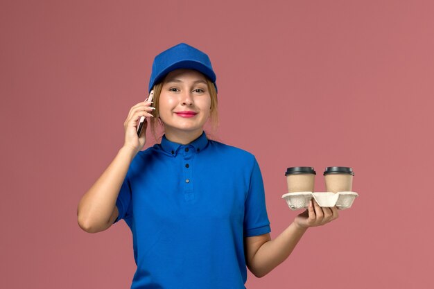 Kurierin in blauer Uniform, die Lieferbecher Kaffee hält und am Telefon auf rosa, Dienstarbeiterjobuniformlieferung spricht