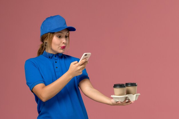 Kurierin in blauer Uniform, die Lieferbecher Kaffee hält und ihr Telefon auf rosa, Dienstjobuniformlieferung verwendet