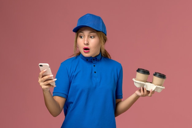 Kurierin in blauer Uniform, die Lieferbecher Kaffee hält und ihr Telefon mit schockiertem Ausdruck auf rosa, Dienstuniformzustellung verwendet