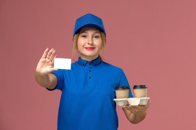 Kurierin in blauer Uniform, die Lieferbecher Kaffee und weiße Karte auf rosa, Dienstuniform-Lieferarbeiter hält