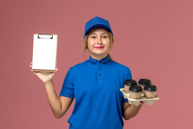 Kurierin in blauer Uniform mit Notizblock zusammen mit braunen Lieferbechern Kaffee auf hellrosa, Dienstuniform Lieferung