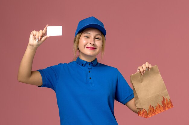 Kurierin in blauer Uniform mit weißer Karte und Lebensmittelpaket auf hellrosa, Dienstuniform-Lieferauftrag