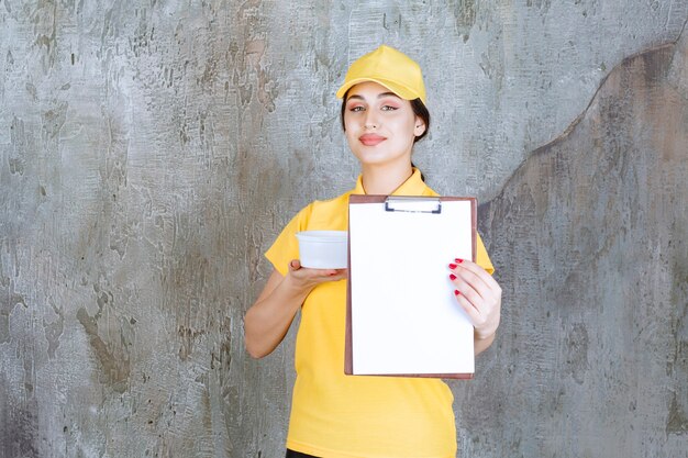 Kurierin in gelber Uniform, die eine Tasse zum Mitnehmen hält und die Aufgabenliste zur Unterschrift vorlegt