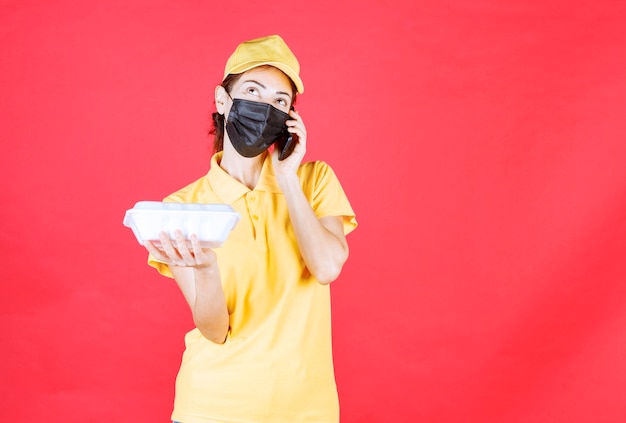 Kurierin in gelber Uniform und schwarzer Maske, die ein Paket zum Mitnehmen hält und Bestellungen per Smartphone entgegennimmt, während sie mit dem Telefon spricht