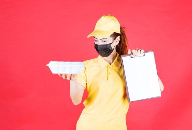 Kurierin in gelber Uniform und schwarzer Maske, die ein Paket zum Mitnehmen hält und die Unterschriftenliste vorlegt