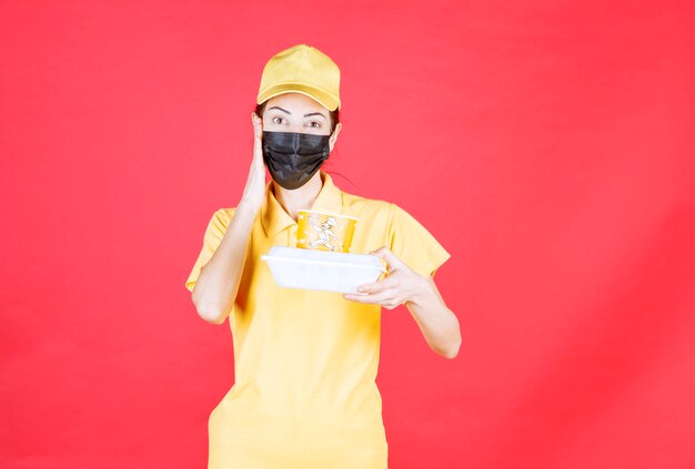 Kurierin in gelber Uniform und schwarzer Maske, die ein Paket zum Mitnehmen hält und verwirrt und nachdenklich aussieht