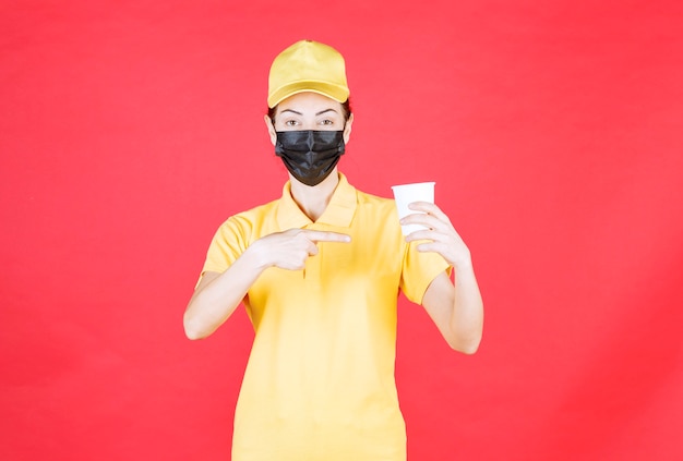 Kurierin in gelber Uniform und schwarzer Maske mit einer Tasse zum Mitnehmen