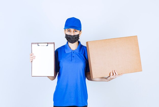 Kurierin in Maske und blauer Uniform, die ein großes Papppaket hält und die Checkliste zur Unterschrift vorlegt