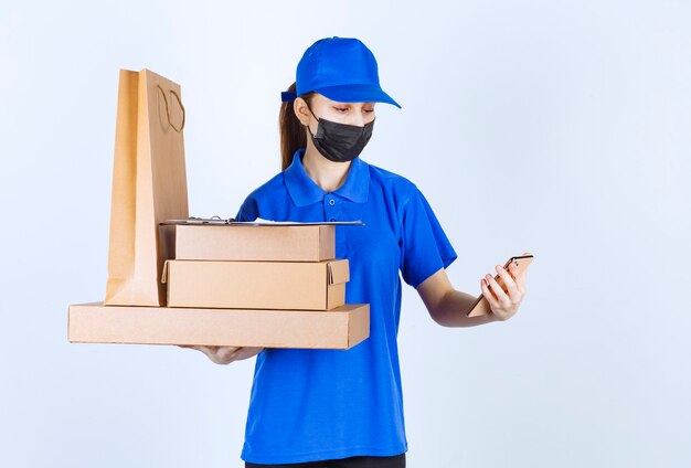 Kurierin in Maske und blauer Uniform, die eine Einkaufstasche aus Karton und mehrere Kisten hält, während sie ihr Selfie macht.