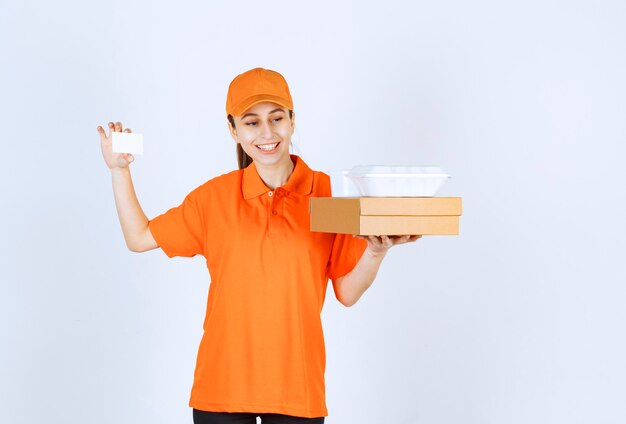 Kurierin in orangefarbener Uniform, die eine Pappschachtel und eine Plastikbox zum Mitnehmen hält, während sie ihre Visitenkarte vorlegt