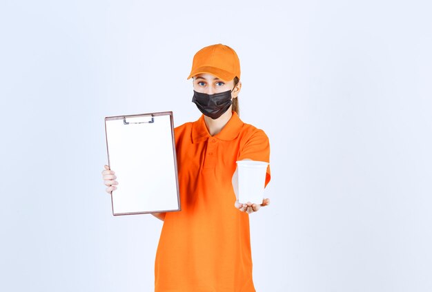 Kurierin in orangefarbener Uniform und schwarzer Maske, die einen Plastikbecher zum Mitnehmen hält und den Unterschriftenrohling zur Übergabe vorlegt