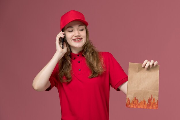 Kurierin in roter Uniform, die braunes Lebensmittelpaket hält, das am Telefon auf dem rosa, einheitlichen Dienstlieferungsjobarbeiter spricht