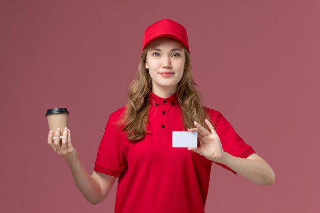 Kurierin in roter Uniform, die Kaffeetasse und Karte auf hellrosa, Arbeitsuniform-Servicearbeiterlieferung hält