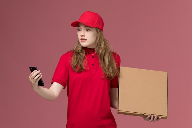 Kurierin in roter Uniform, die Lebensmittelbox hält und ihr Telefon auf rosa, einheitlichem Dienst liefert, der Arbeiter liefert