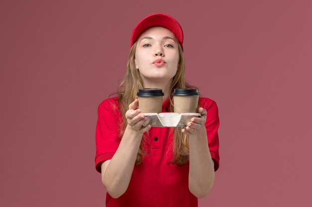 Kurierin in roter Uniform mit braunen Kaffeetassen, die Luftküsse auf rosa, einheitliche Arbeitsvermittlerin senden