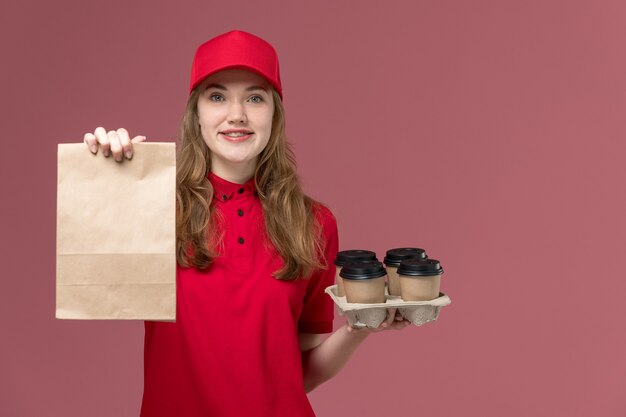 Kurierin in roter Uniform mit Lieferung Lebensmittelpaket mit Kaffeetassen auf hellrosa, Dienstuniform Service Lieferung