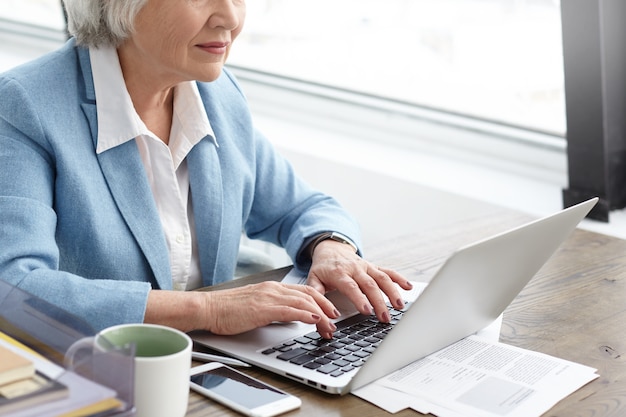 Kurzer Schuss einer älteren Geschäftsfrau mit grauem Haar und faltigen Händen, die auf Laptop schreiben, während sie in ihrem Büro arbeiten. Stilvolle reife kaukasische Frau, die blauen Anzug mit Geräten für die Arbeit trägt